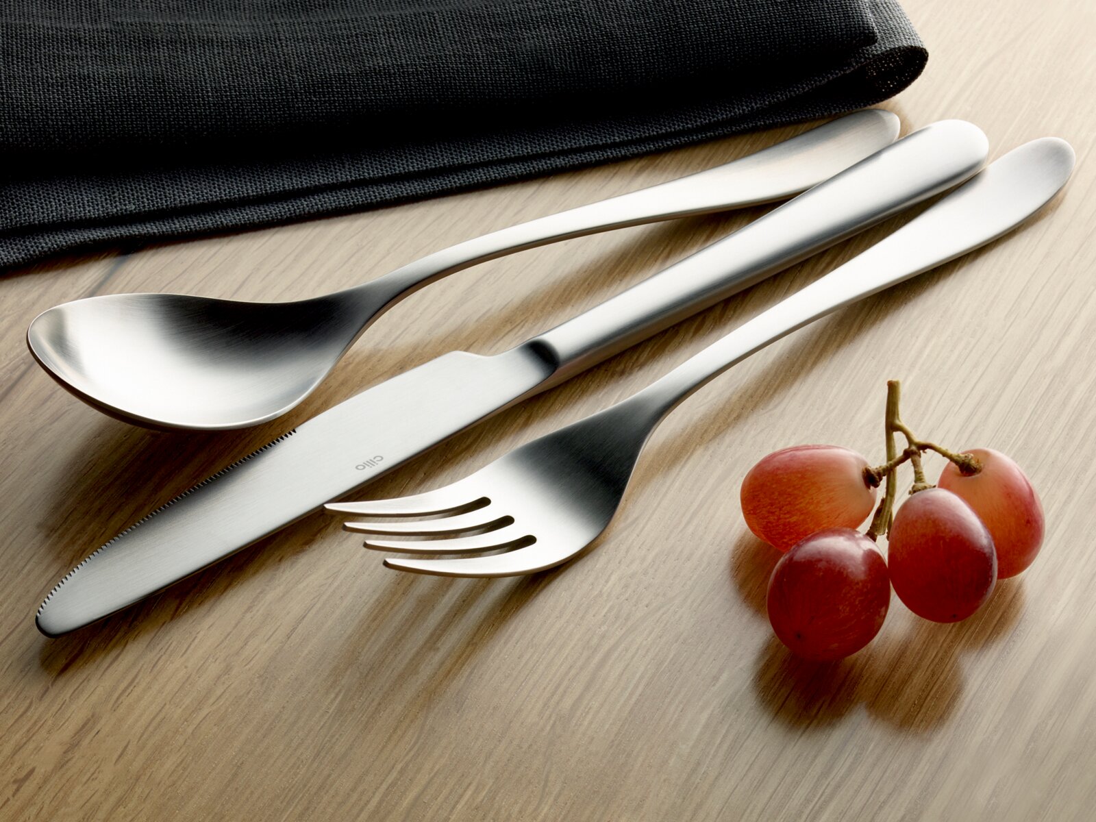 Elegantes Besteck-Set mit Löffel, Messer und Gabel aus Edelstahl, auf einem Holztisch mit Trauben und Stoffserviette, seitliche Perspektive.