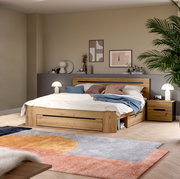 Modernes Holzbett in Eiche-Optik mit integriertem Stauraum und Schublade, weißer Bettwäsche und blauen Akzentkissen im Schlafzimmer; Perspektive: frontale Ansicht leicht von links auf das Bett