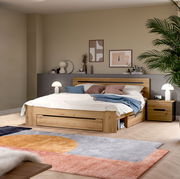 Modernes Holzbett in Eiche-Optik mit integriertem Stauraum und Schublade, weißer Bettwäsche und blauen Akzentkissen im Schlafzimmer; Perspektive: frontale Ansicht leicht von links auf das Bett