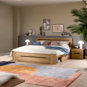 Modernes Holzbett in Eiche-Optik mit integriertem Stauraum und Schublade, weißer Bettwäsche und blauen Akzentkissen im Schlafzimmer; Perspektive: frontale Ansicht leicht von links auf das Bett