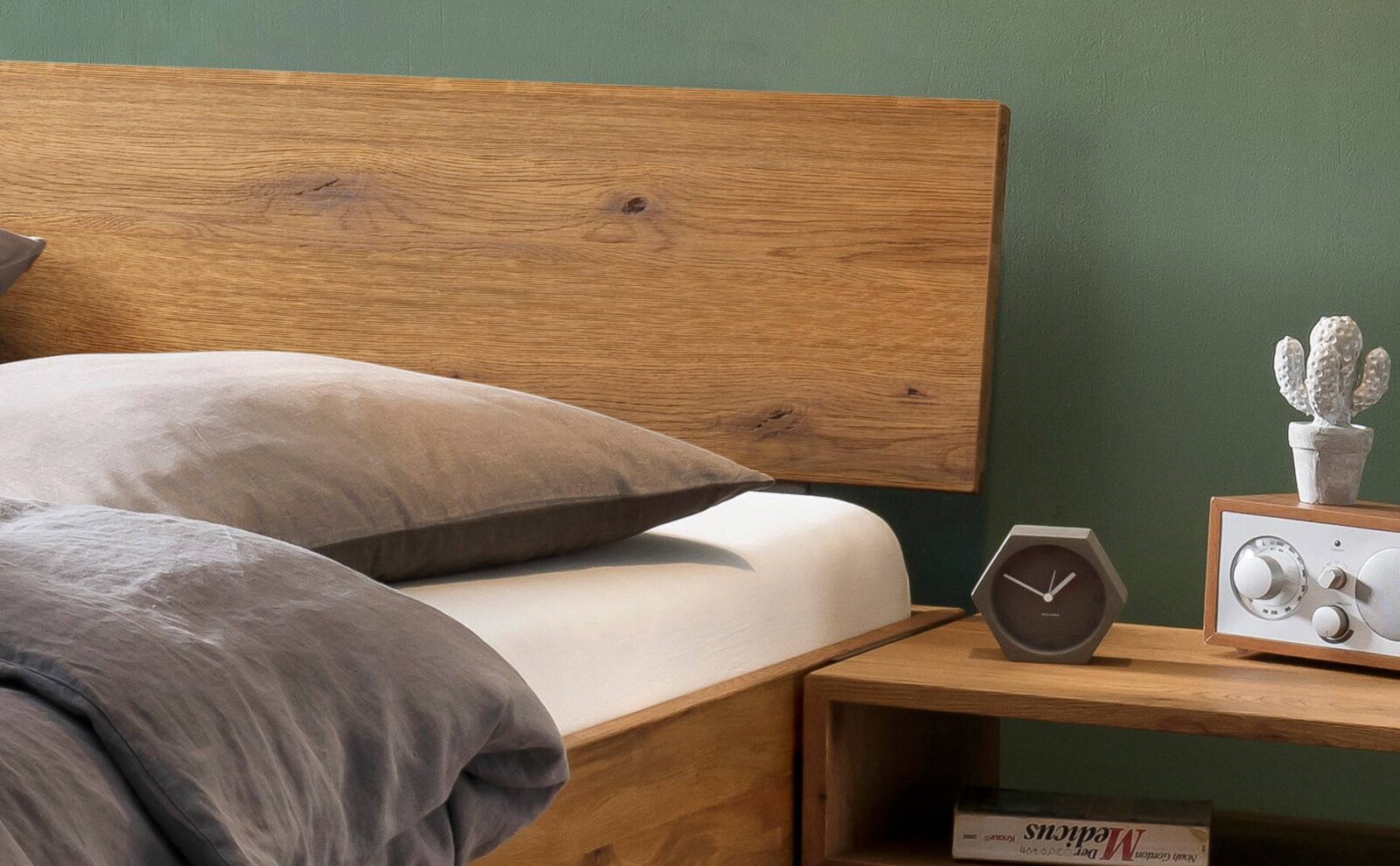 Modernes Holzbett mit breitem Kopfteil und grauer Bettwäsche neben Nachttisch mit Uhr, Radio und kleiner Kaktus-Deko, seitliche Perspektive in Nahaufnahme