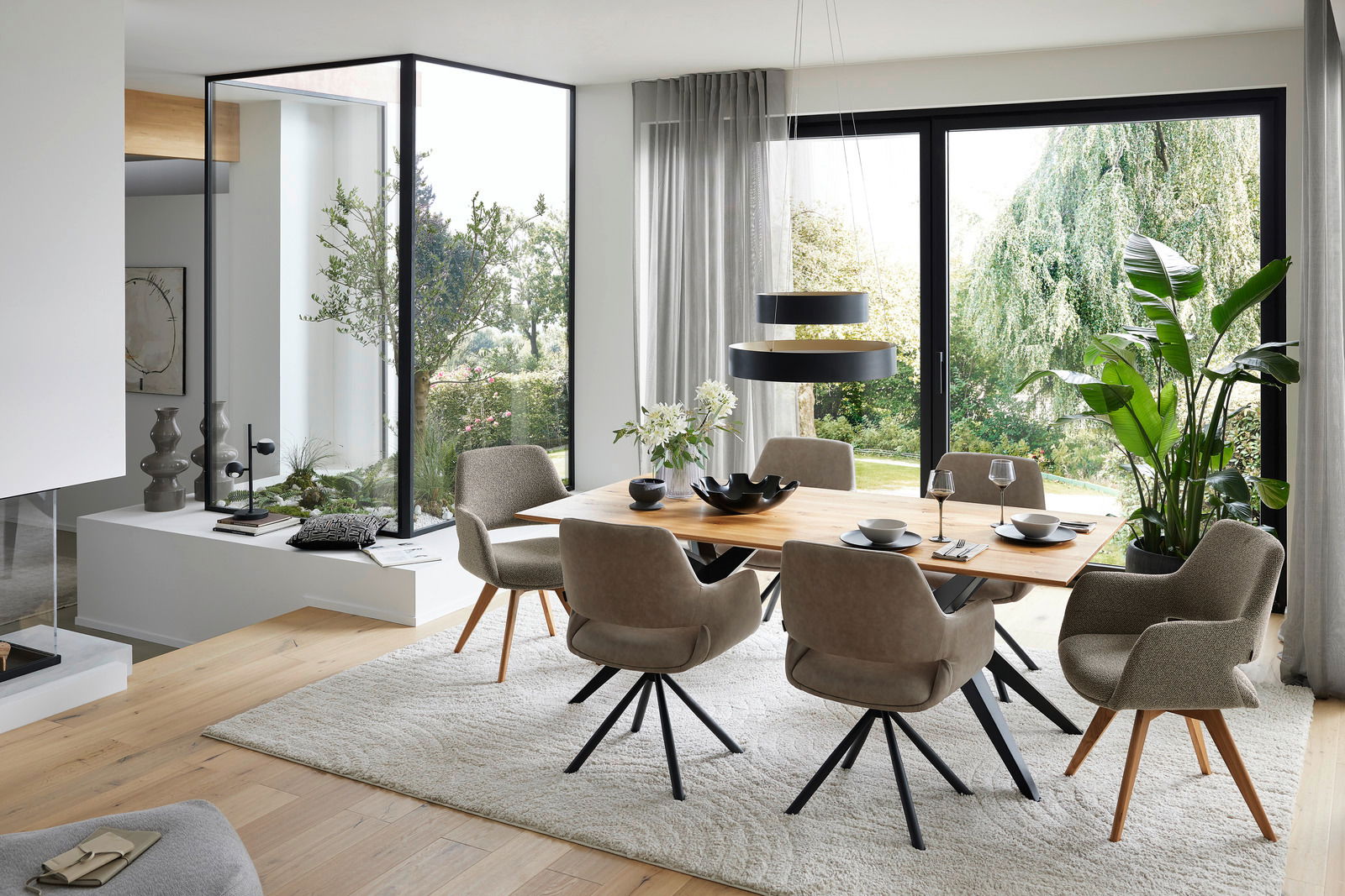 Musterring Armlehnstuhl MR TARIO Moderner Essbereich mit großem Holztisch und bequemen Armlehnstühlen, umgeben von großen Fenstern mit Blick ins Grüne. Perspektive: seitlich.