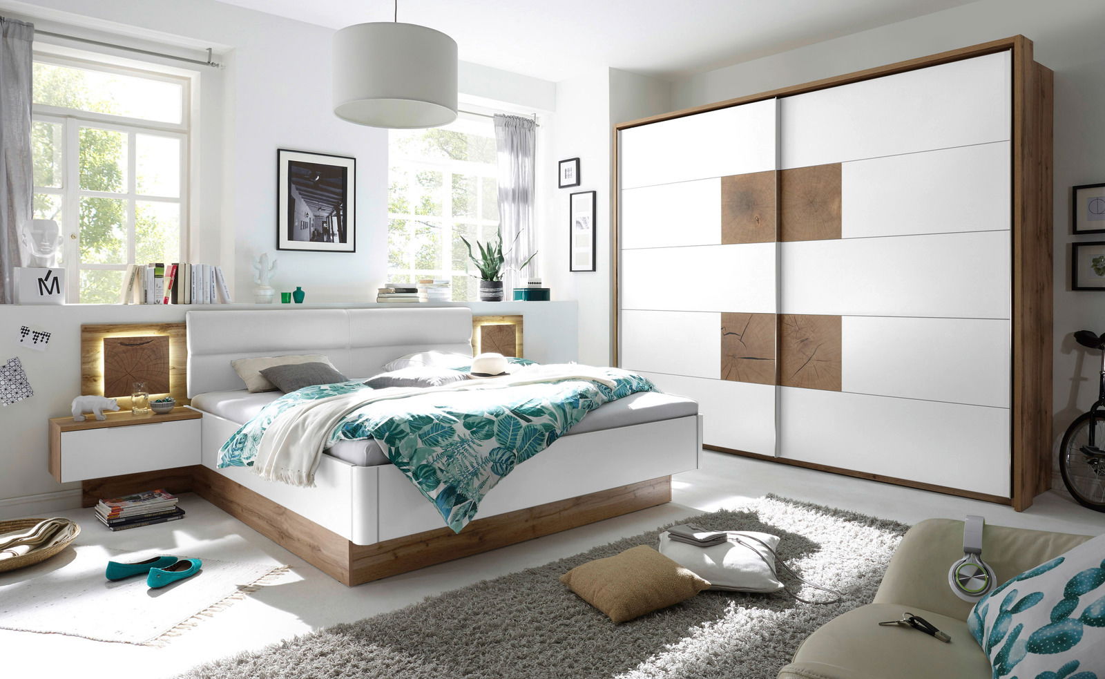 Moderne Schlafzimmermöbel mit einem weißen Schwebetürenschrank und einem Bett mit Holz- und Weißakzenten, seitliche Perspektive.