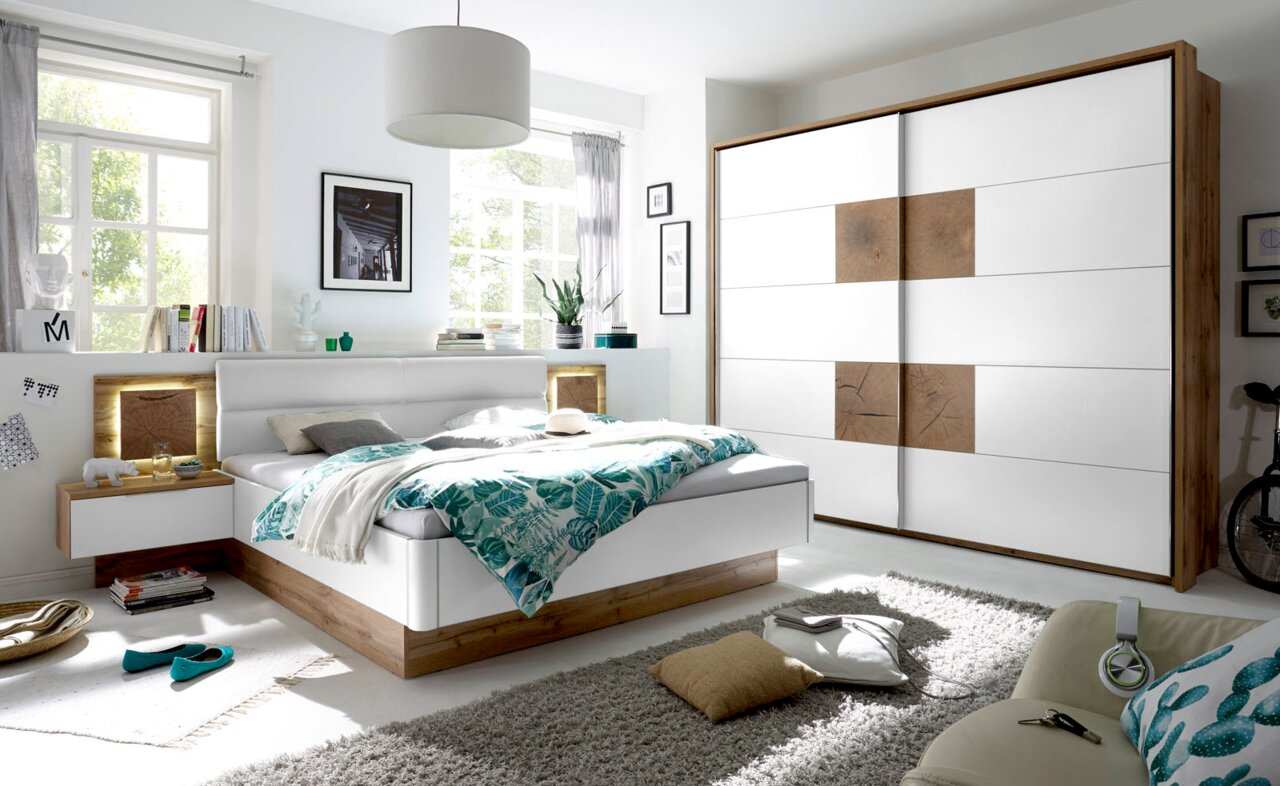 Moderne Schlafzimmermöbel mit einem weißen Schwebetürenschrank und einem Bett mit Holz- und Weißakzenten, seitliche Perspektive.