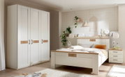 Helles Schlafzimmer-Set im Landhausstil mit weißem Bett, Nachttisch und großem Kleiderschrank, Akzenten in Holzoptik und schwarzem Griffdesign; Perspektive: frontale Raumansicht leicht schräg von links.