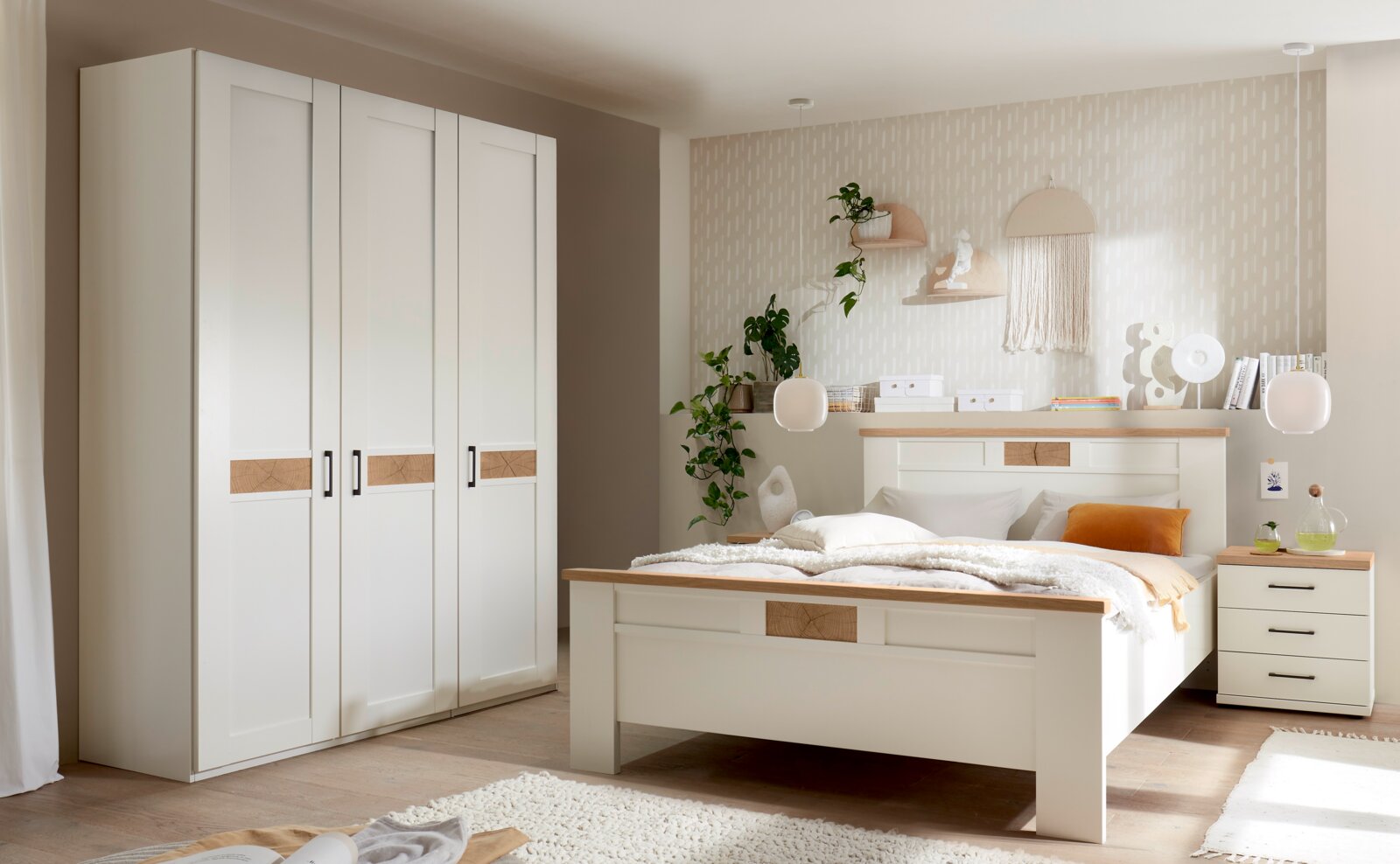 Helles Schlafzimmer-Set im Landhausstil mit weißem Bett, Nachttisch und großem Kleiderschrank, Akzenten in Holzoptik und schwarzem Griffdesign; Perspektive: frontale Raumansicht leicht schräg von links.