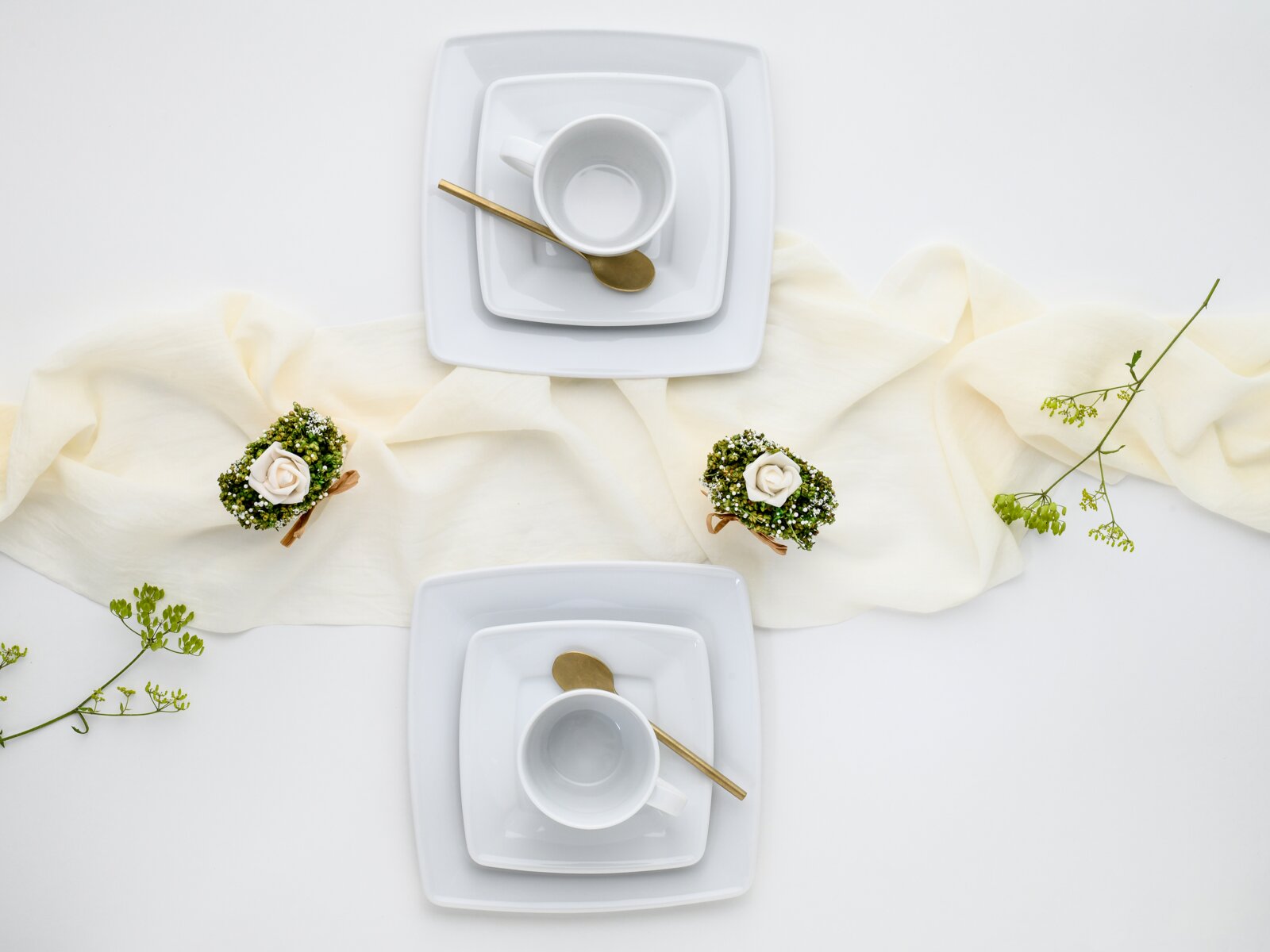 Draufsicht auf zwei weiße, quadratische Porzellan-Tassen mit Untertassen und goldfarbenen Löffeln, dekoriert mit hellem Stoffband und kleinen Blumenarrangements auf weißem Hintergrund