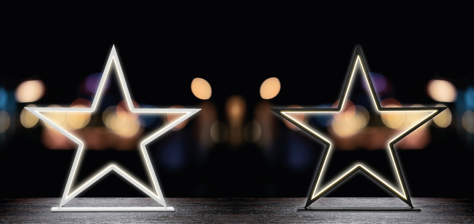 Zwei leuchtende LED-Sterne auf einem Tisch vor einem unscharfen, dunklen Hintergrund, frontal betrachtet.