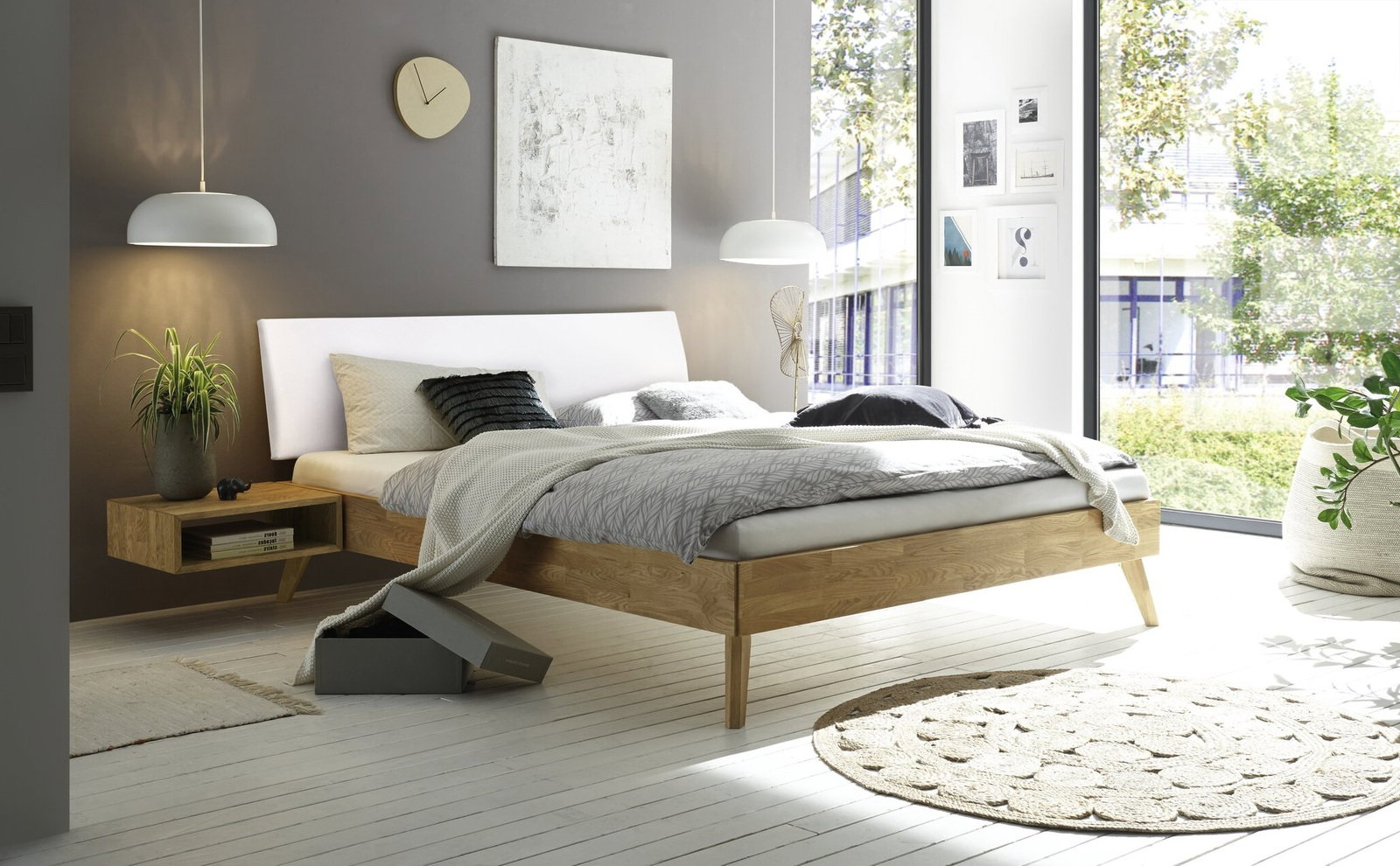 Modernes Bettgestell aus Holz mit weißem Kopfteil, seitliche Perspektive, umgeben von stilvoller Dekoration in einem hellen Raum mit großen Fenstern.