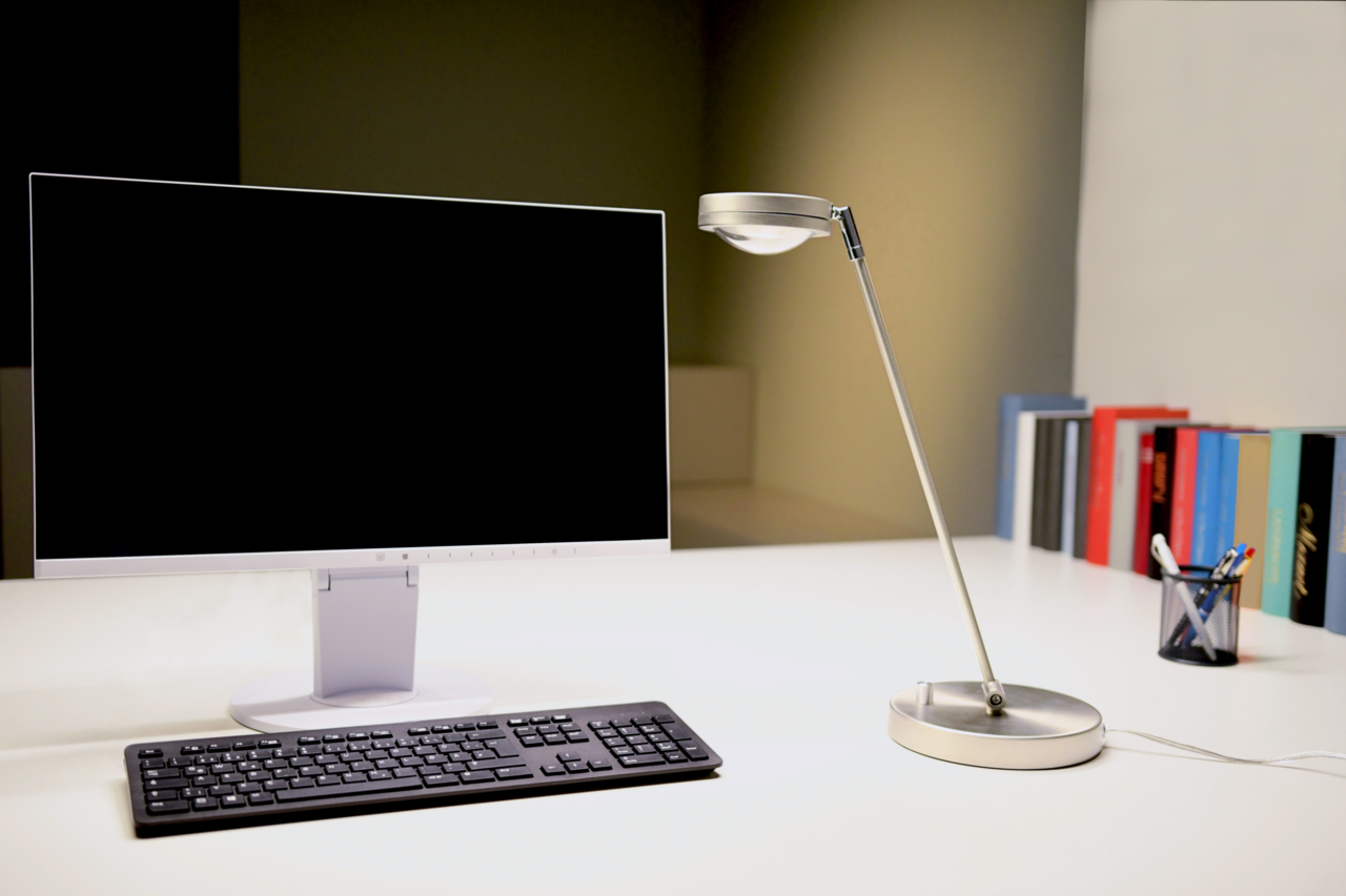 Moderne LED Tischleuchte aus Metall in seitlicher Perspektive auf einem Schreibtisch neben einem Monitor und einer Tastatur.