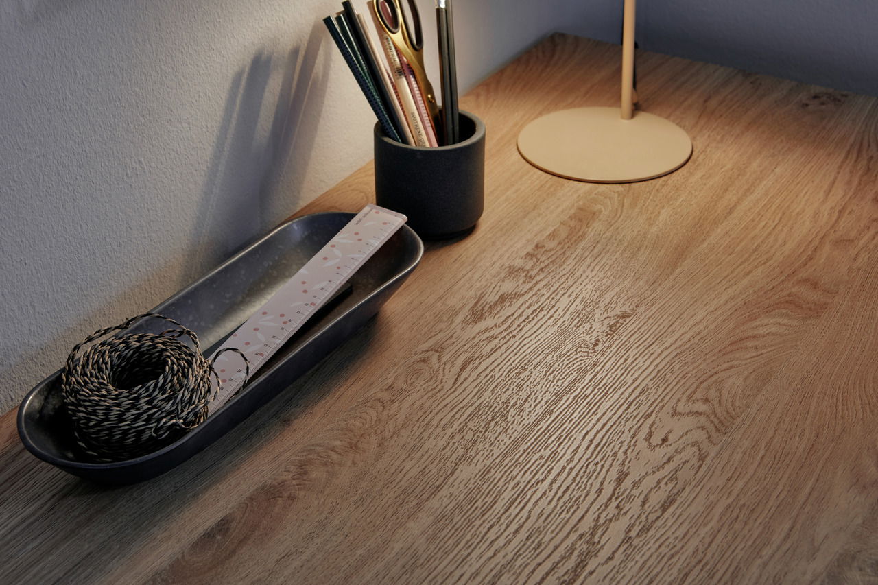 Nahaufnahme eines Eck-Schreibtisches aus Holz mit Schreibtischutensilien und Lampe, von oben betrachtet.