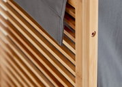 Detailaufnahme aus schräger Seitenperspektive: Holzrahmen mit Lamellen und grauem Stoffbezug, Nahansicht der Kante und Befestigungsöffnung.