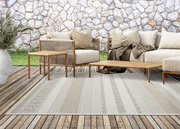 Beiger Outdoor-Teppich mit dezentem Streifen- und Strukturmuster auf Holzterrasse vor Steinmauer, umgeben von Lounge-Möbeln; Perspektive: frontale, leicht erhöhte Ansicht.