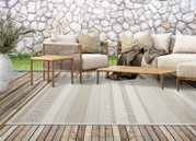 Beige buitentapijt met een subtiel streep- en structuurpatroon op een houten terras voor een stenen muur, omringd door loungemeubilair; perspectief: frontaal, licht verhoogd.