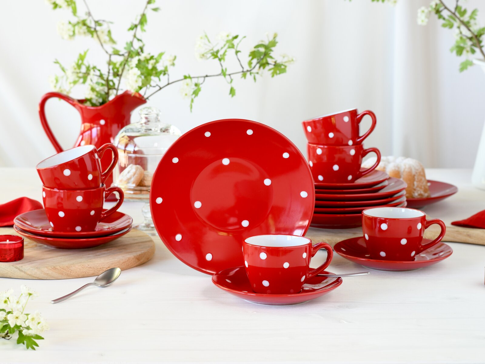 Rotes Keramik-Kaffeeservice mit weißen Punkten (Tassen, Untertassen und Teller) auf weißem Tisch arrangiert, frontal in leichter Schrägansicht fotografiert; im Hintergrund rote Kanne, Glasglocke mit Gebäck und Frühlingszweige.
