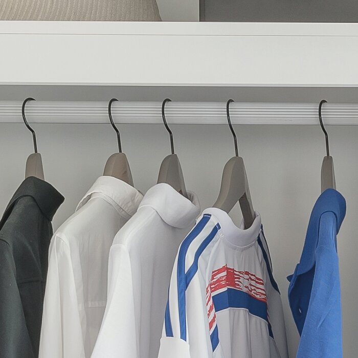 Kleding aan hangers in een open kledingkast: witte overhemden en een wit sportshirt met blauw-rode strepen naast een blauwe trui, frontaal aanzicht op ooghoogte.