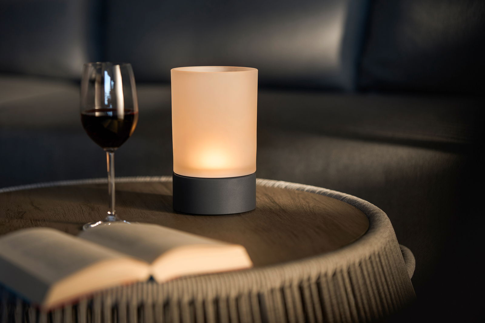 Stimmungsvolle Akku-Tischleuchte auf einem Tisch, neben einem Weinglas und einem Buch, aus seitlicher Perspektive fotografiert.