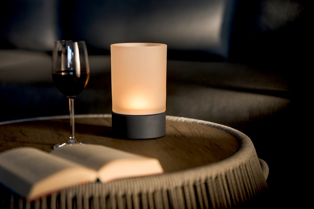 Stimmungsvolle Akku-Tischleuchte auf einem Tisch, neben einem Weinglas und einem Buch, aus seitlicher Perspektive fotografiert.