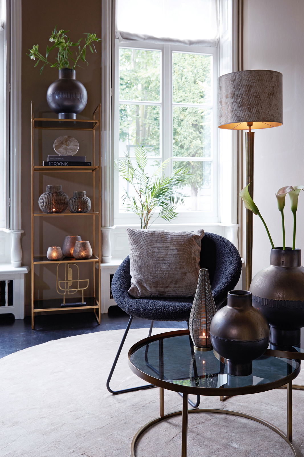 Light&Living Uhr MORENO Elegante Wohnzimmerdekoration mit einer braunen Marmor-Uhr auf einem goldenen Regal, umgeben von stilvollen Vasen und Pflanzen, im Tageslicht eines großen Fensters.