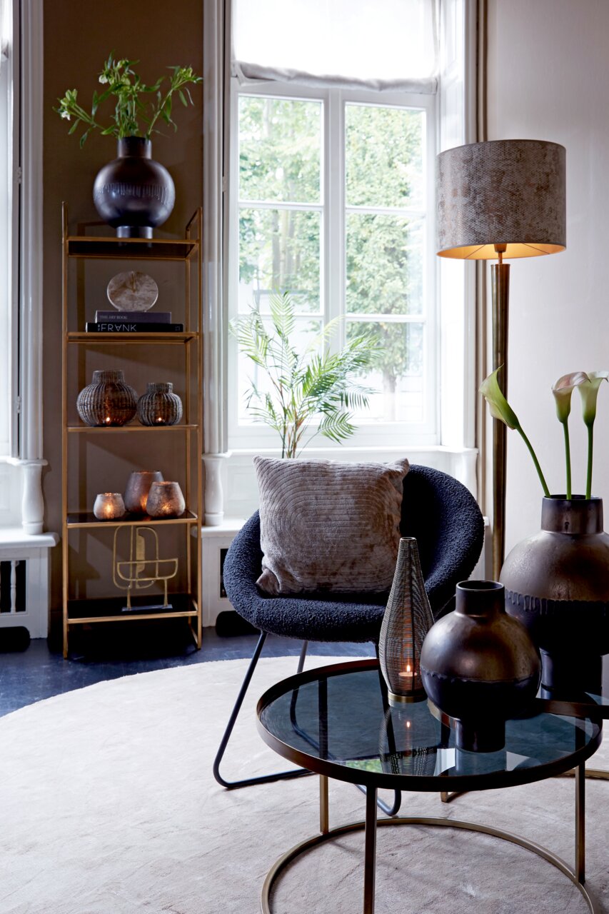 Light&Living Uhr MORENO Elegante Wohnzimmerdekoration mit einer braunen Marmor-Uhr auf einem goldenen Regal, umgeben von stilvollen Vasen und Pflanzen, im Tageslicht eines großen Fensters.