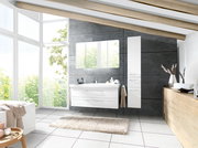Modernes Badezimmer mit weißem Waschtisch mit zwei Schubladen, passendem Spiegelschrank und hohem Hochschrank vor grauer Betonwand, fotografiert in frontaler Perspektive mit leichter Weitwinkelansicht.