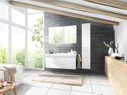 Modernes Badezimmer mit weißem Waschtisch mit zwei Schubladen, passendem Spiegelschrank und hohem Hochschrank vor grauer Betonwand, fotografiert in frontaler Perspektive mit leichter Weitwinkelansicht.