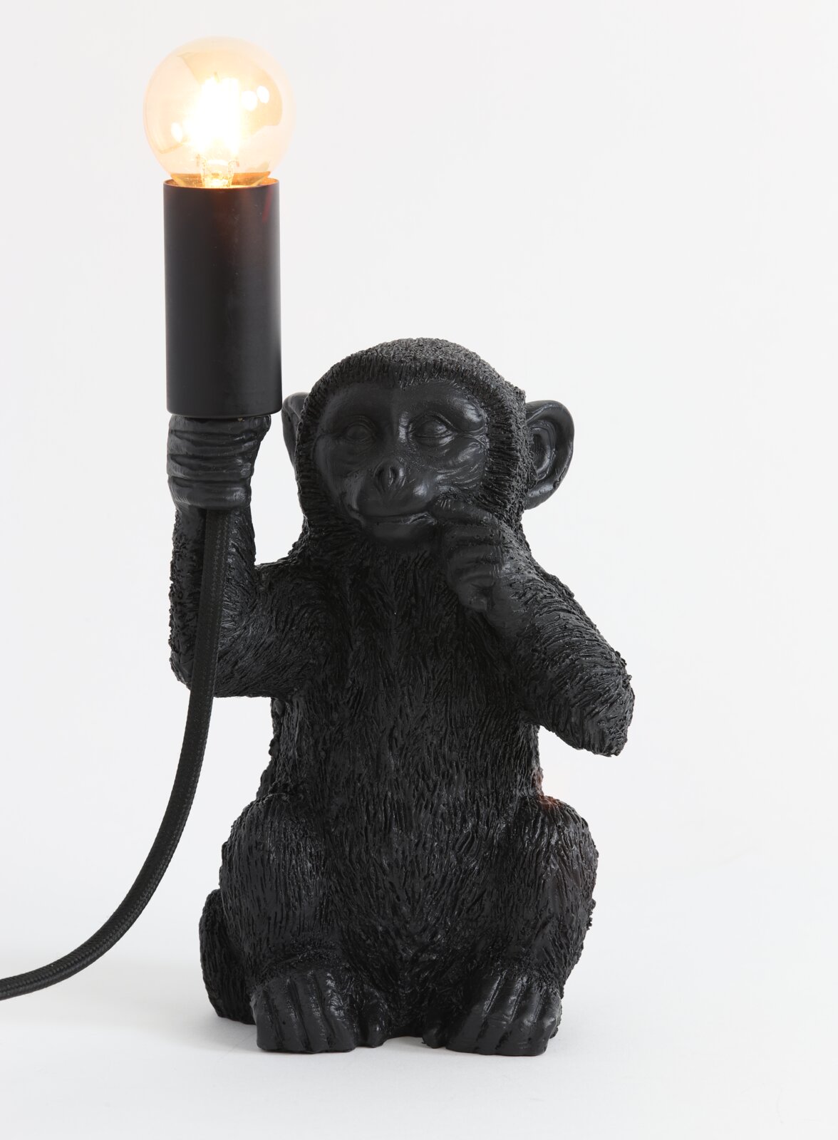 Zwarte tafellamp in de vorm van een aap die een gloeilamp in zijn hand houdt, gezien van voren.