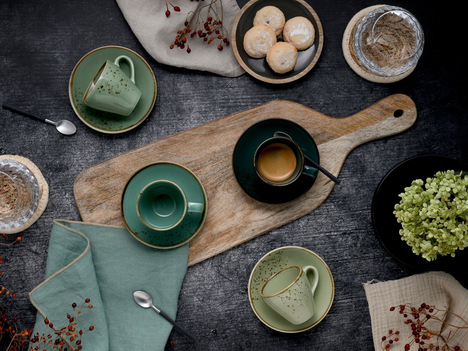 Draufsicht auf ein rustikales Kaffee-Arrangement mit grün gesprenkelten Tassen und Untertassen auf Holzbrett, Espresso, Löffeln, Gebäck und dekorativen Beeren auf dunklem Tisch