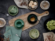 Draufsicht auf ein rustikales Kaffee-Arrangement mit grün gesprenkelten Tassen und Untertassen auf Holzbrett, Espresso, Löffeln, Gebäck und dekorativen Beeren auf dunklem Tisch
