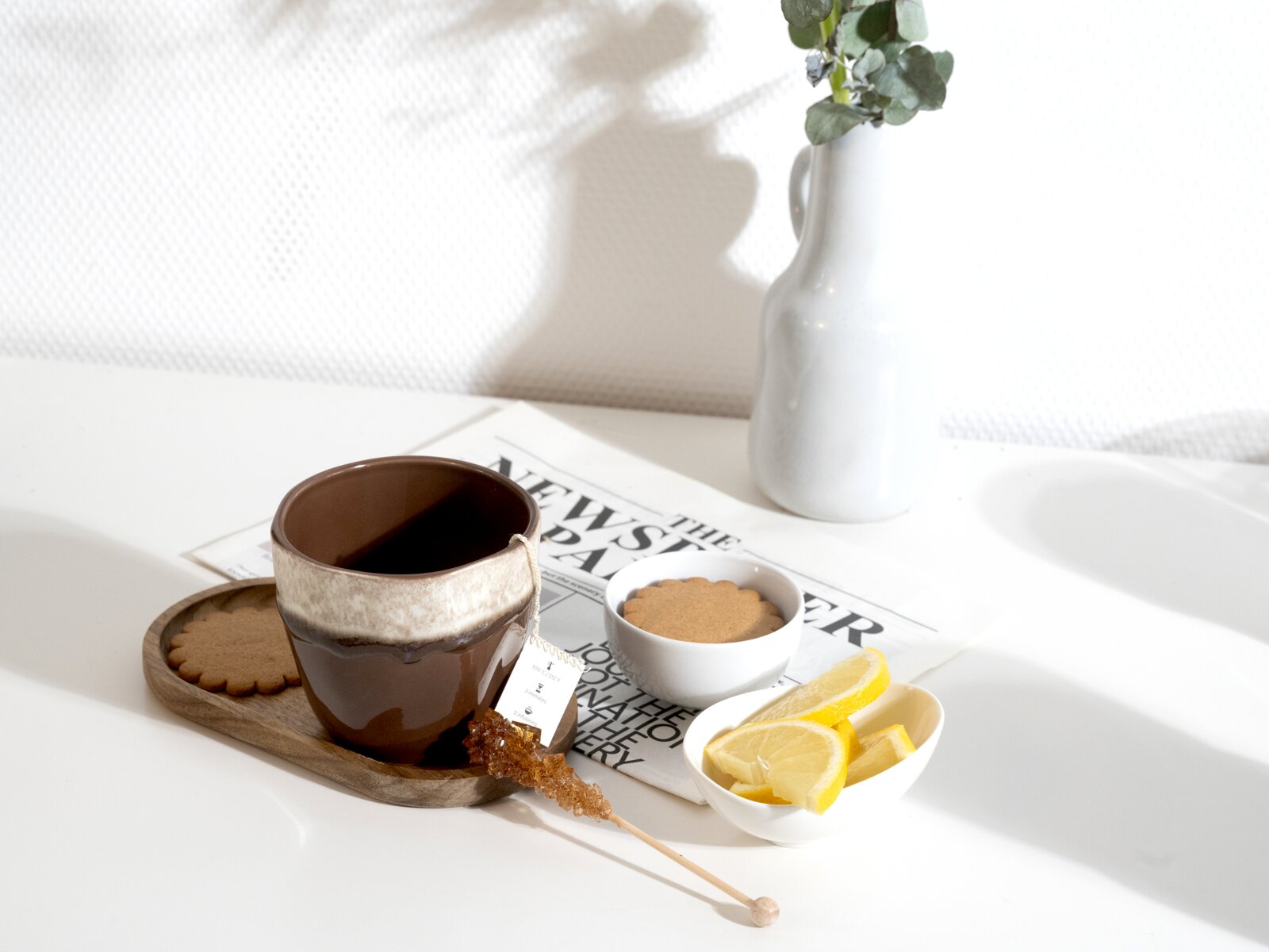 Braune Keramiktasse mit Tee auf Holztablett, dazu Kekse, Zitronenscheiben und Kandiszucker auf hellem Tisch neben Zeitung und weißer Vase, aufgenommen aus leicht erhöhter schräger Perspektive.