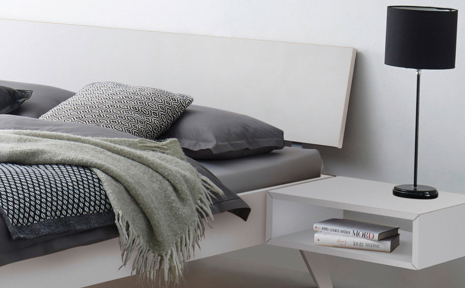 Modernes Bettgestell mit grauer Bettwäsche, Kissen und Decke, seitliche Perspektive mit Nachttisch und Lampe.