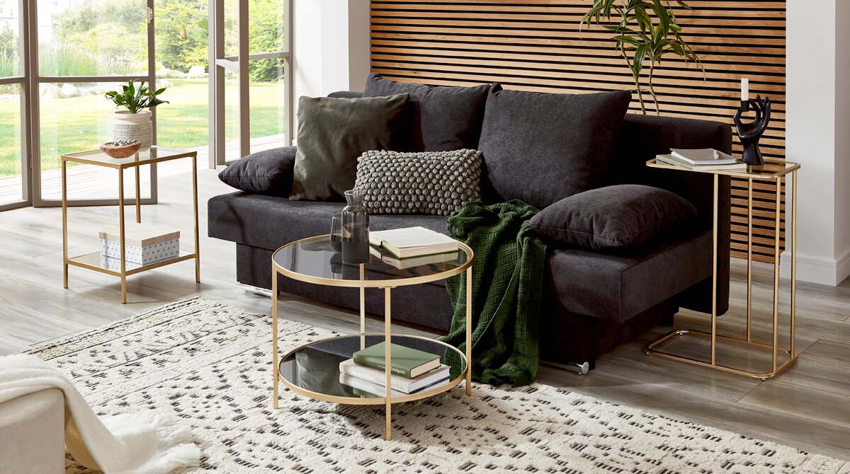 Zurbrüggen Beistelltisch Modernes Wohnzimmer mit dunkelgrauem Sofa, runden Glas-Couchtischen und goldfarbenen Beistelltischen vor Holzlamellenwand, aufgenommen in leicht schräger Frontansicht.