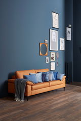 Ein elegantes, dreisitziges Sofa in einem warmen Braunton, dekoriert mit blauen Kissen und einer Decke, vor einer stilvoll gestalteten Wand mit Bildern. Perspektive von vorne links.