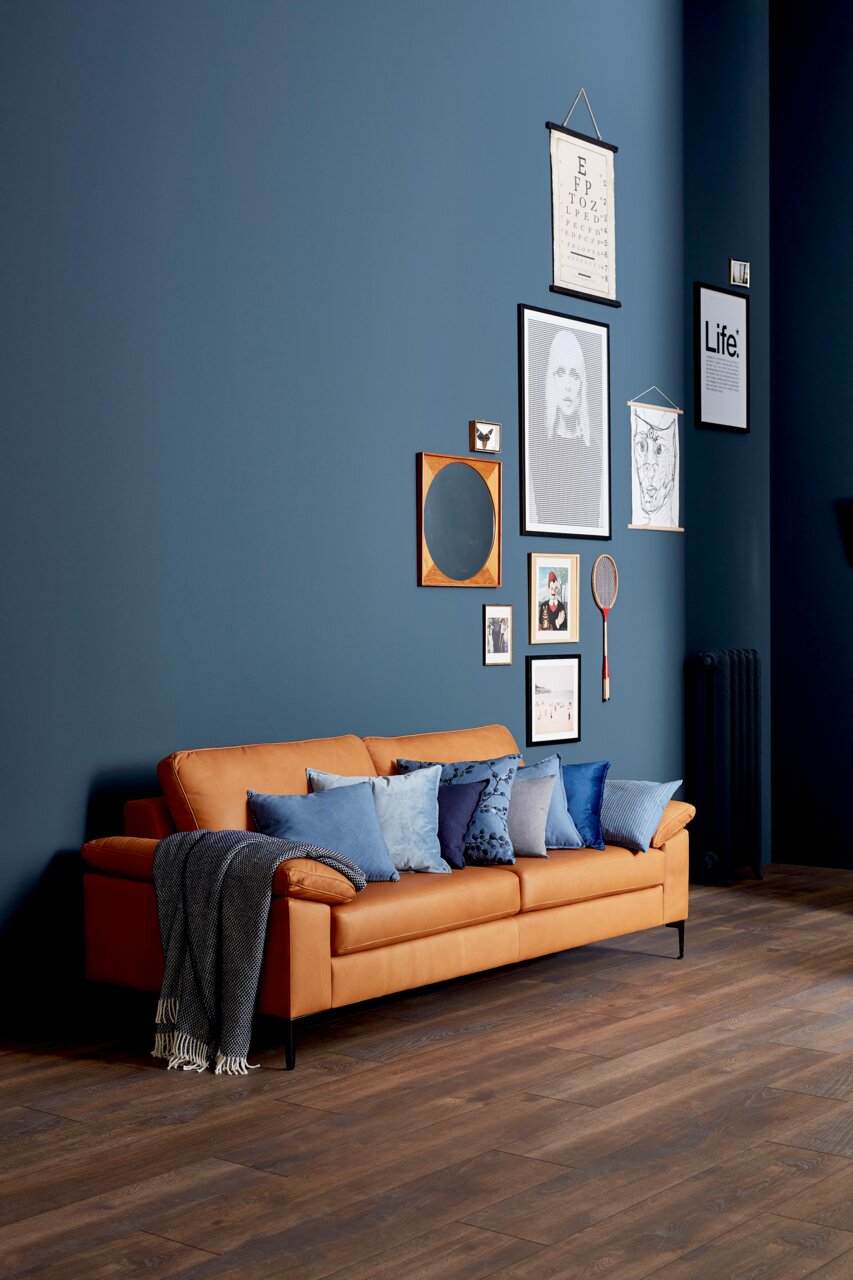 Ein elegantes, dreisitziges Sofa in einem warmen Braunton, dekoriert mit blauen Kissen und einer Decke, vor einer stilvoll gestalteten Wand mit Bildern. Perspektive von vorne links.