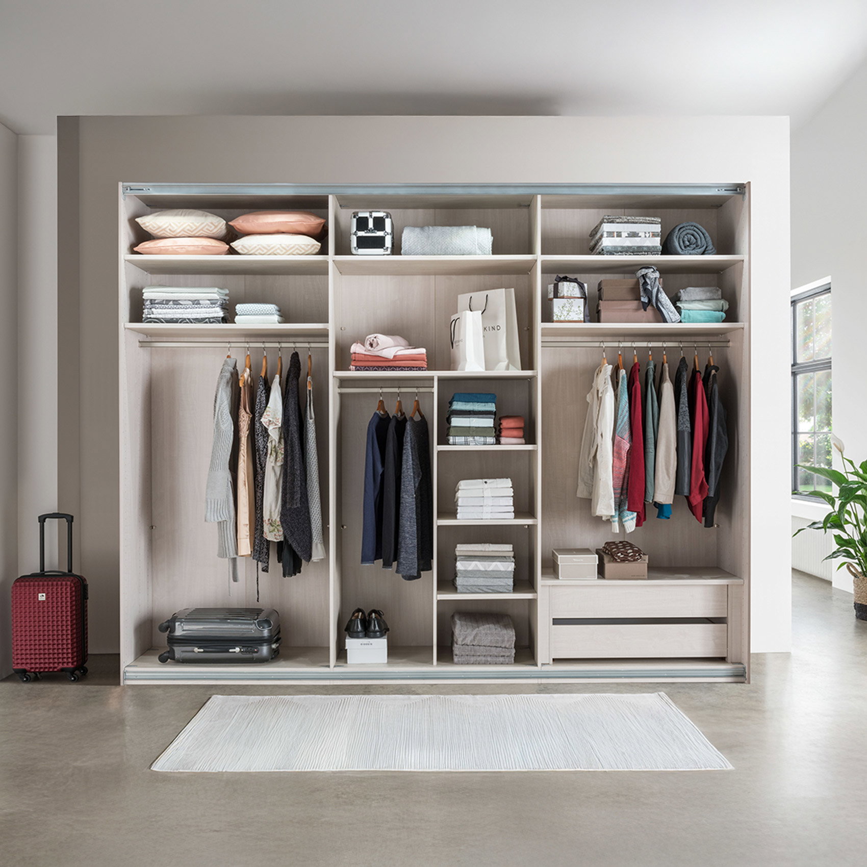 Großer offener Kleiderschrank mit Fächern, Kleiderstangen und Schubladen, ordentlich gefüllt mit Kleidung, Kissen und Boxen, daneben ein Koffer