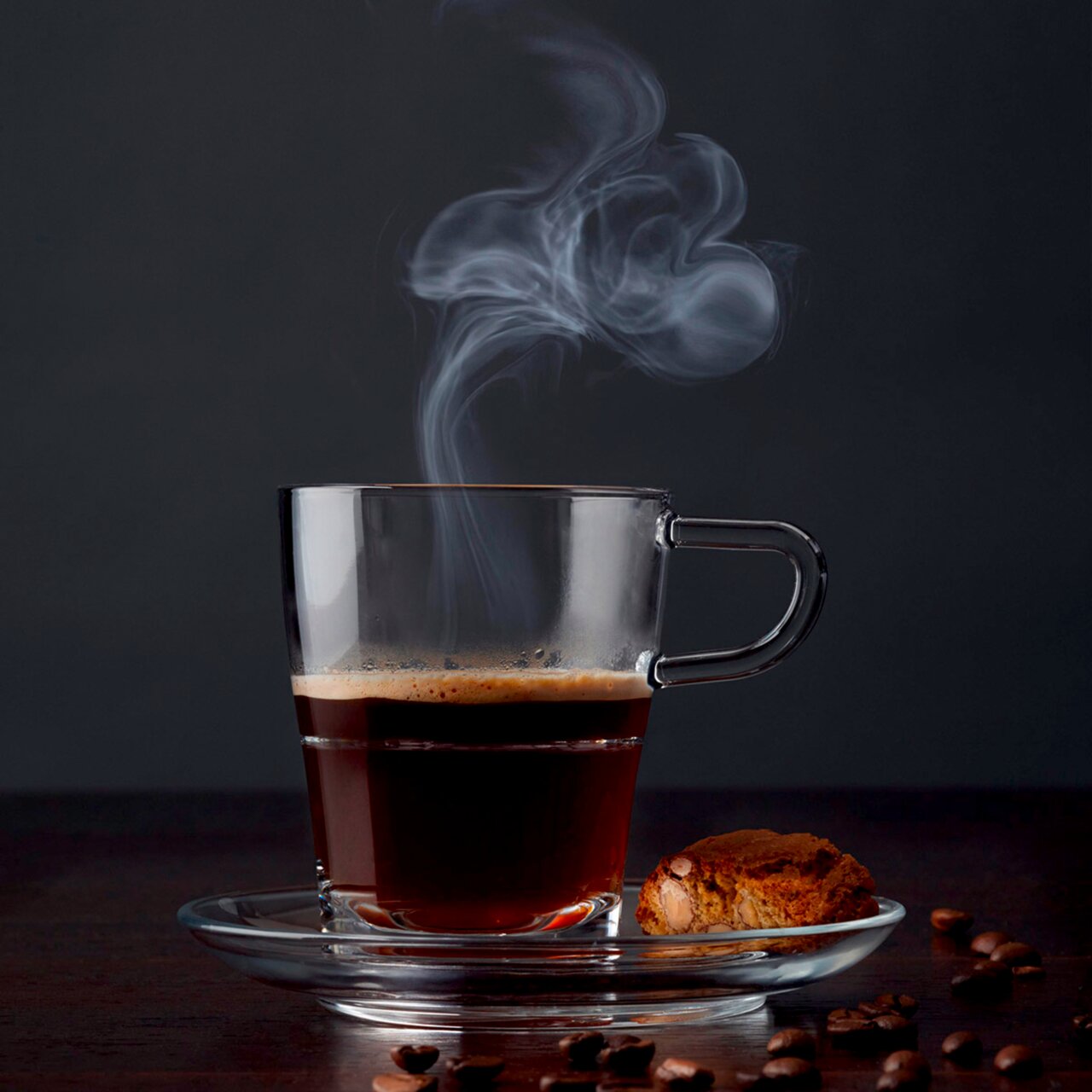 Transparente Espressotasse Senso 70ml mit dampfendem Espresso, von der Seite fotografiert, auf einem Unterteller mit einem Keks und Kaffeebohnen.