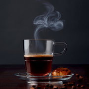 Transparente Espressotasse Senso 70ml mit dampfendem Espresso, von der Seite fotografiert, auf einem Unterteller mit einem Keks und Kaffeebohnen.