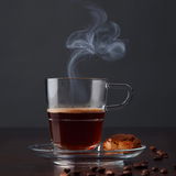 Transparente Espressotasse Senso 70ml mit dampfendem Espresso, von der Seite fotografiert, auf einem Unterteller mit einem Keks und Kaffeebohnen.