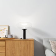 Moderne Tischleuchte mit klarem Glas und schwarzem Sockel auf einem Holzschrank, seitliche Perspektive.