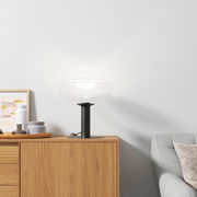 Moderne Tischleuchte mit klarem Glas und schwarzem Sockel auf einem Holzschrank, seitliche Perspektive.