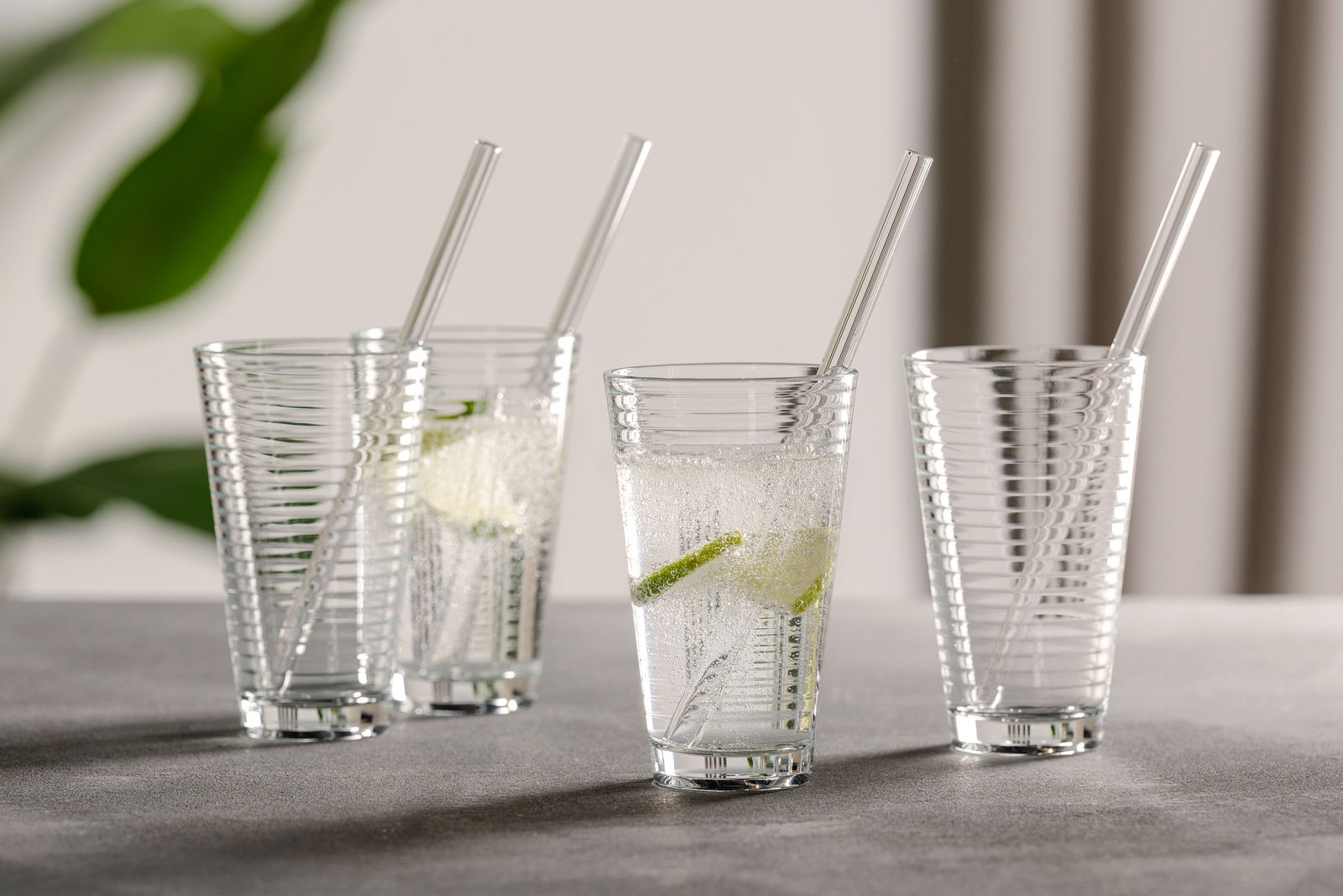 Vier durchsichtige Becher mit Spiraldesign und Trinkhalm, gefüllt mit sprudelndem Wasser und Limettenscheiben, auf einer grauen Oberfläche. Seitliche Perspektive.