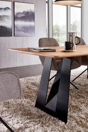 Moderne eettafel met een lichtgekleurd houten blad en een zwart V-vormig metalen onderstel, zijaanzicht in de woon-/eetkamer met stoelen, een kopje en een kan op de tafel