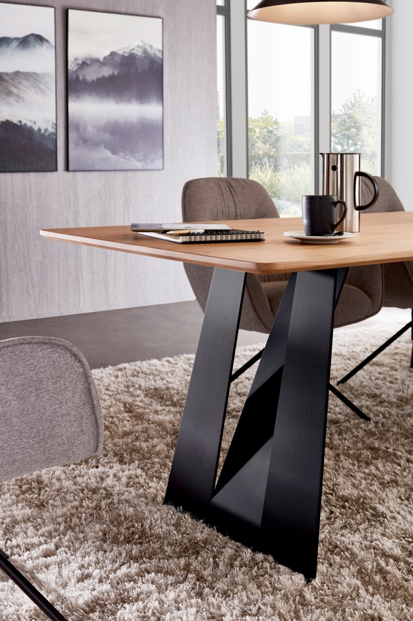 Moderne eettafel met een lichtgekleurd houten blad en een zwart V-vormig metalen onderstel, zijaanzicht in de woon-/eetkamer met stoelen, een kopje en een kan op de tafel