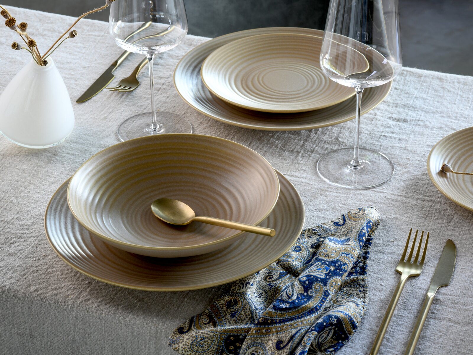 Elegantes Steingut-Geschirrset in Beige mit feinen Rillen, kombiniert mit goldfarbenem Besteck und Weingläsern auf Leinentischdecke, perspektivisch in schräger Draufsicht aufgenommen