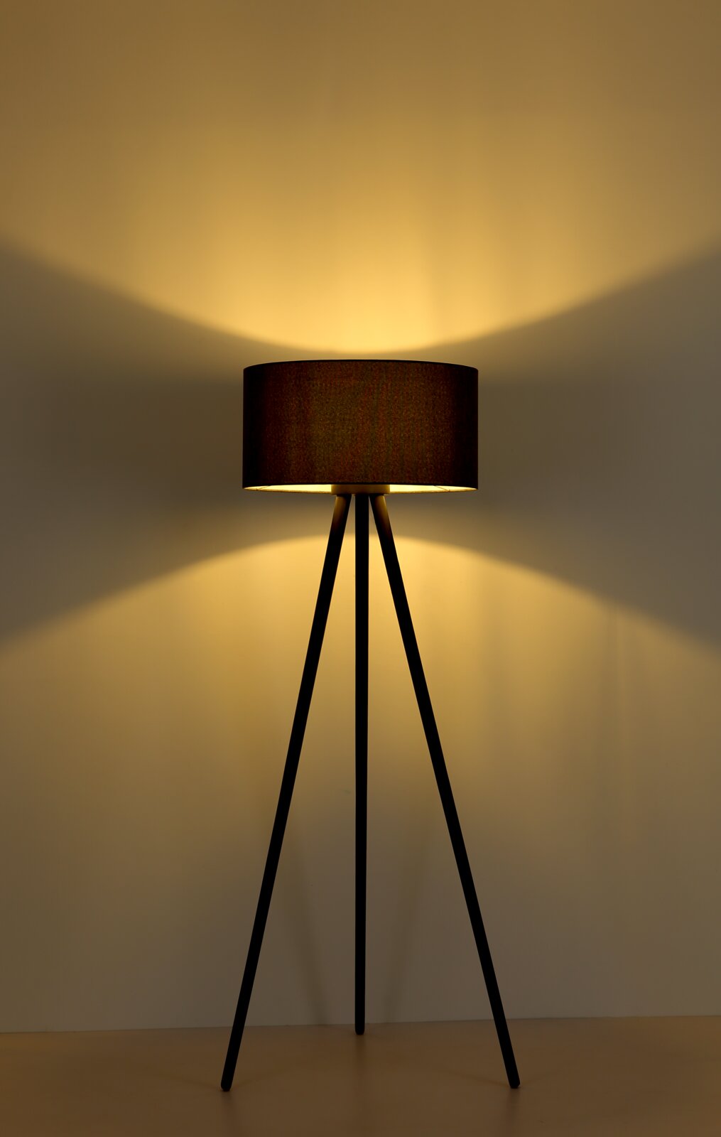 Moderne Stehlampe mit schwarzem Dreibein-Gestell und dunkelbraunem zylindrischem Stoffschirm, eingeschaltet mit warmem Lichtkegel an der Wand, frontale Perspektive.