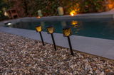 Drei Solar-Erdspieße GRETITA in einem Garten, die bei Dämmerung entlang eines Pools leuchten, seitliche Perspektive.