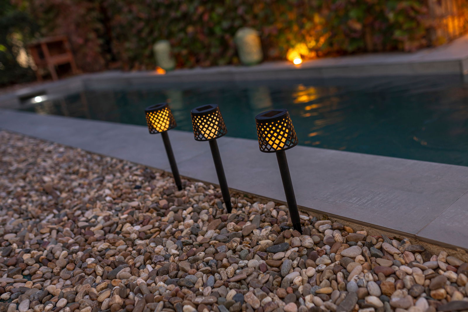 Drei Solar-Erdspieße GRETITA in einem Garten, die bei Dämmerung entlang eines Pools leuchten, seitliche Perspektive.
