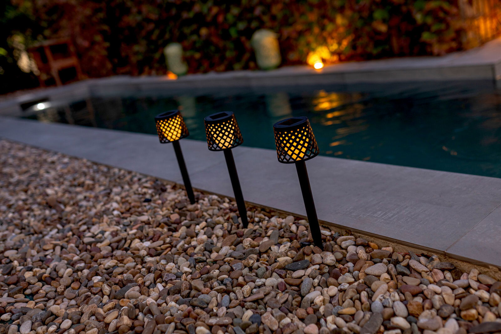 Drei Solar-Erdspieße GRETITA in einem Garten, die bei Dämmerung entlang eines Pools leuchten, seitliche Perspektive.