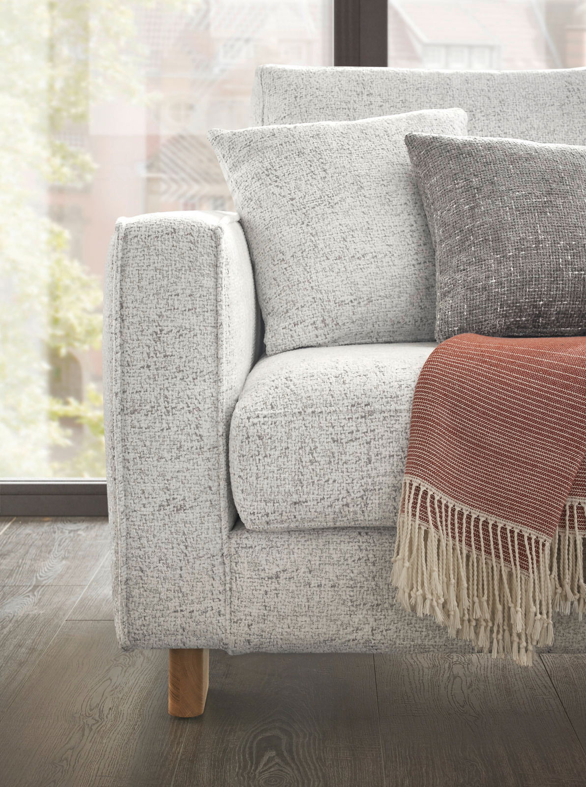 Nahaufnahme eines modernen Ecksofas mit hellgrauem Stoffbezug, dekoriert mit Kissen und einer roten Decke, aus seitlicher Perspektive.