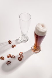 Zwei Weizenbiergläser, eines leer und eines mit dunklem Bier gefüllt, von oben fotografiert, umgeben von kleinen Brezeln auf einem weißen Hintergrund.